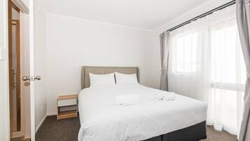 Una cama bien hecha con sábanas blancas, una mesita de noche de madera y una ventana con cortinas traslúcidas.