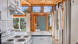 A modern kitchen with a central island, a stove, and a dining area with a view of palm trees. - Dry Dock: Your Cozy Beachside Retreat at 612 Eastern Lake Rd (Seagrove Beach)