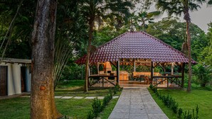 Un gazebo con tetto in tegole, circondato da vegetazione rigogliosa e un vialetto lastricato che vi conduce.