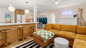 Een moderne keuken met houten kastjes, een wit aanrecht en een barkruk. Er staat een bank met een geruite plaid en een girafdecoratie.
