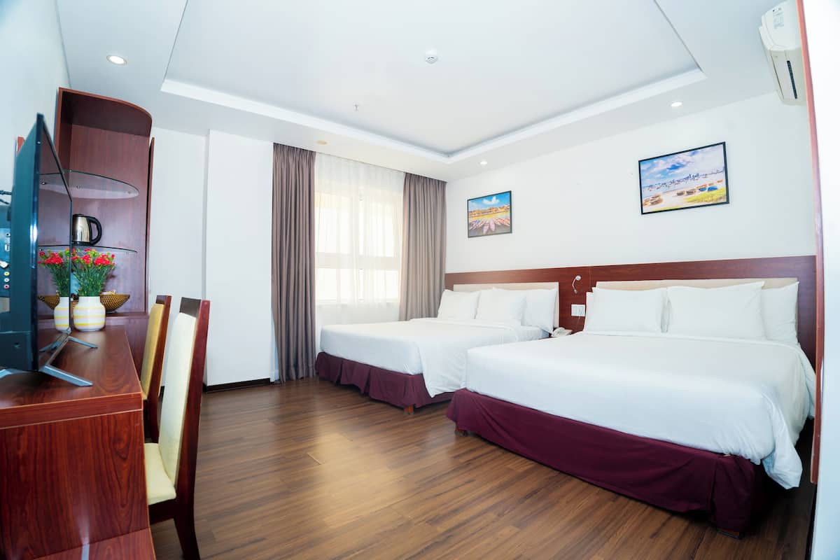 A hotel room with two beds, wooden flooring, a desk with a TV, and a window with curtains.