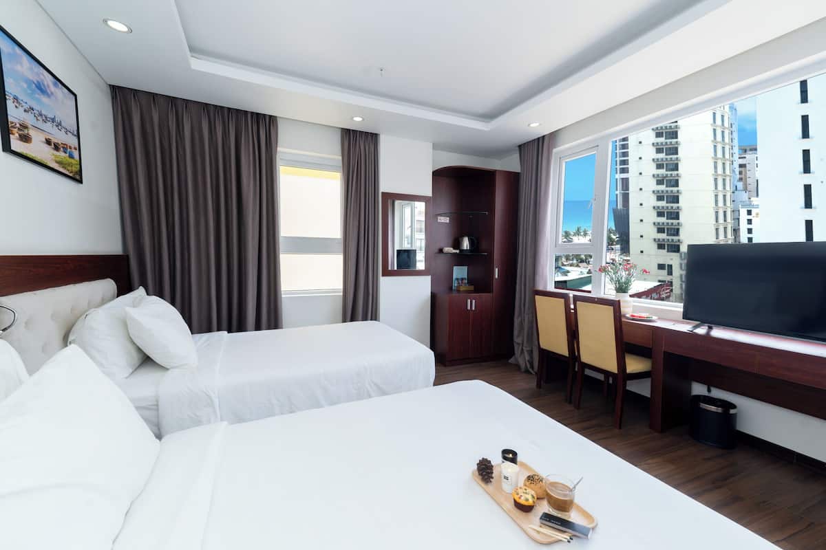 A hotel room with two beds, a desk with a TV, a window with a view of buildings, and a tray with food items.