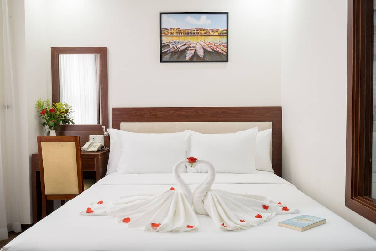 A hotel room with a neatly made bed, a wooden headboard, a bedside table with a telephone, a chair, and a framed picture on the wall.