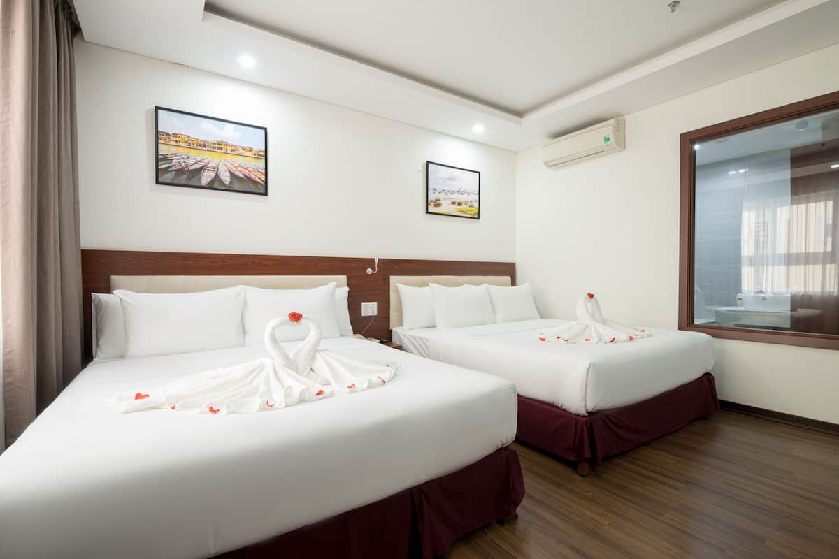 A hotel room with two beds, each with a white bedspread and red skirted base. There are two framed pictures on the wall, an air conditioning unit, and a window with a view of the outside.