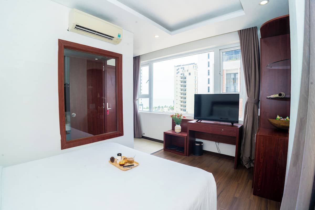 A hotel room with a bed, a TV, a desk, a window with a view of buildings, and an air conditioning unit.