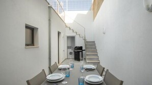 Uma área de refeições ao ar livre e estreita, com uma mesa posta para seis pessoas, uma churrasqueira e uma escadaria que leva a um andar superior.