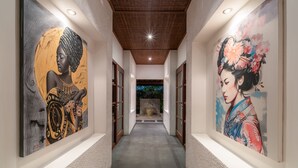 A hallway with two large paintings on the walls, a wooden ceiling, and a view of a garden through an open door at the end. - Villa Claudia  (Kerobokan)
