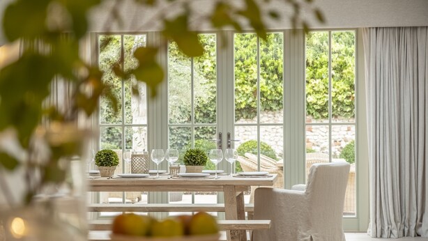 Un comedor con una mesa preparada para cuatro, un frutero y vistas al exterior con vegetación.