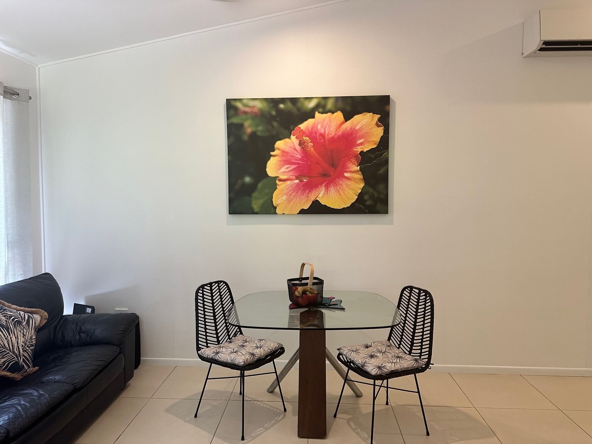 A dining area with a glass table, two chairs, and a hibiscus flower painting on the wall.