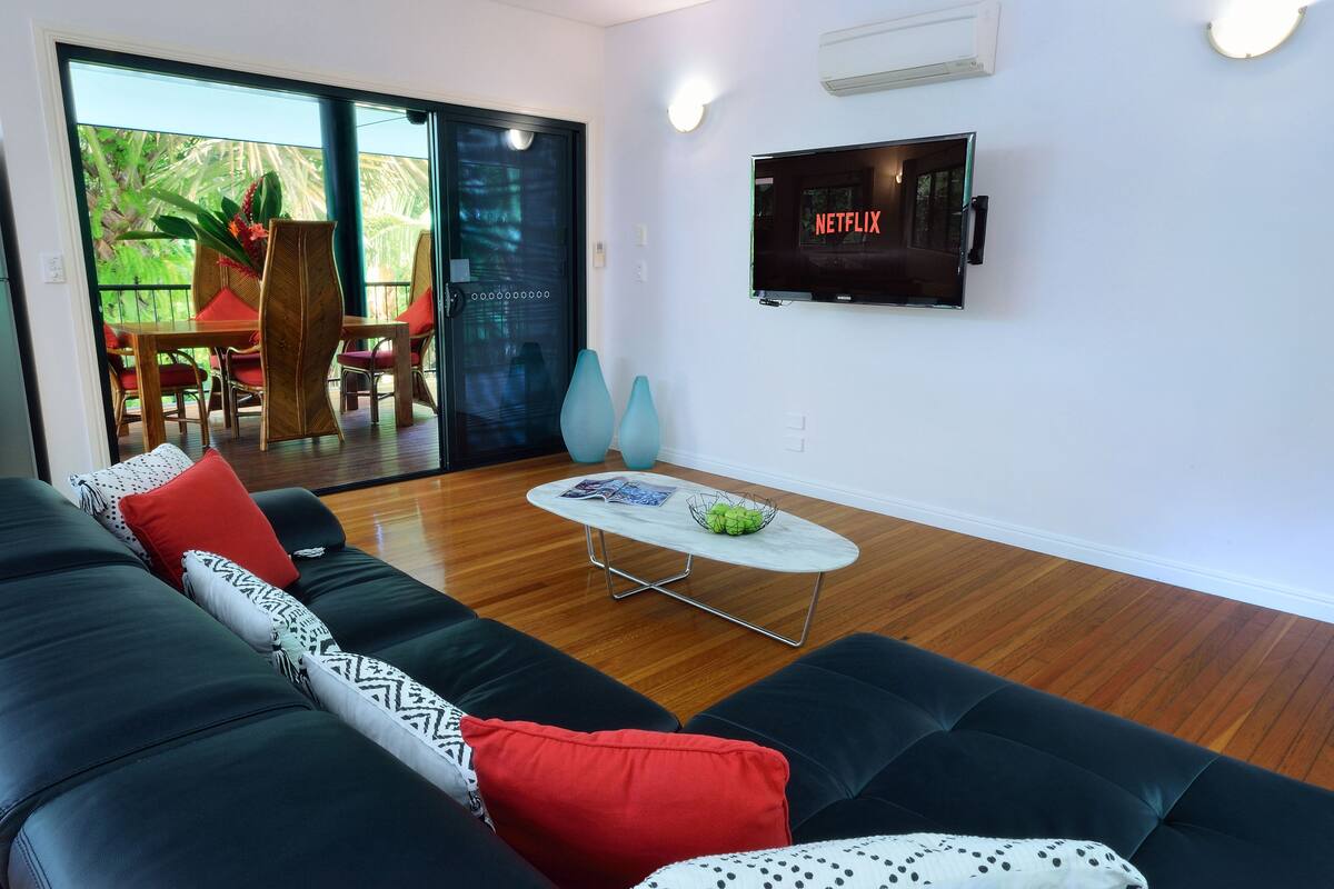 Ein modernes Wohnzimmer mit einer schwarzen Ledersofa, einer Glastür zu einem Außenbereich, einem flachbildfernseher an der Wand und einem kleinen runden Couchtisch.