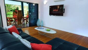 A modern living room with a black leather sofa, a glass door leading to an outdoor area, a flat-screen TV mounted on the wall, and a small round coffee table.