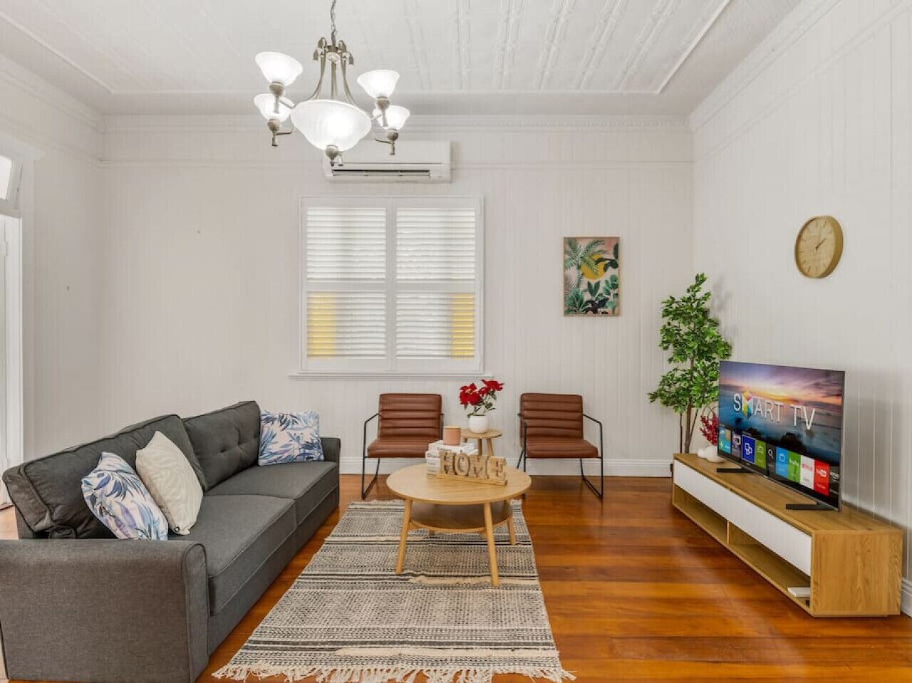 Ein Wohnzimmer mit einem grauen Sofa, zwei braunen Sesseln, einem Couchtisch mit einer floralen Dekoration, einem Fernseher auf einem Holzständer und einer Pflanze.