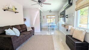 A living room with a brown sofa, a coffee table, a dining area with a table and chairs, a ceiling fan, and a large window with blinds. - Peaceful Beach Home Fully Equipped! (Sarasota)