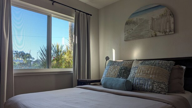 A bedroom with a large bed, a window with curtains, a wall-mounted painting, and a nightstand with a lamp. - Peaceful Beach Home Fully Equipped! (Sarasota)