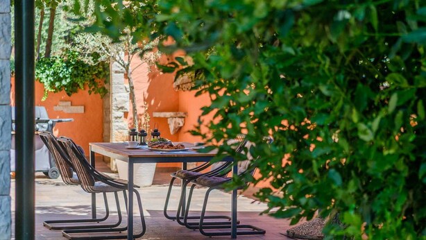 Une terrasse extérieure avec une table et des chaises, entourée de verdure et d’un mur en pierre.