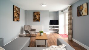 Un salon moderne avec un canapé, un fauteuil et une table basse. On y trouve un téléviseur posé sur un meuble et une fenêtre avec des rideaux.