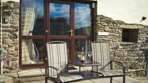 A stone cottage with a large glass door, two striped chairs, a small table, and a bottle of wine.