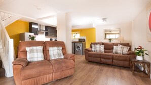 A living room with two brown sofas, a wooden floor, a kitchen in the background, and a staircase.