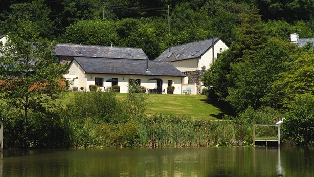 A two-story house with a sloped roof, surrounded by lush greenery and a body of water. - Smithy Cottage at Lower Aylescott (West Down)