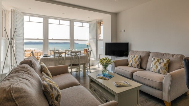 A spacious living area with a large window overlooking a beach, a flat-screen TV, and a comfortable seating arrangement. - 8 Hartland, sleeps 6 with sea view (Esplande, Woolacombe)