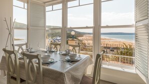 A dining area with a table set for four, large windows with a beach view, and a balcony with a railing. - 8 Hartland, sleeps 6 with sea view (Esplande, Woolacombe)