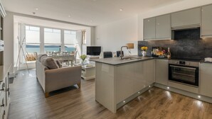 A modern kitchen with a central island, a dining area with a view, and a living space with a sofa and TV. - 8 Hartland, sleeps 6 with sea view (Esplande, Woolacombe)