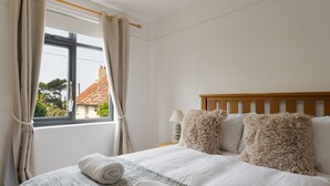 A bedroom with a wooden headboard, two fluffy pillows, a bedside lamp, and a view of a house through a window with curtains.