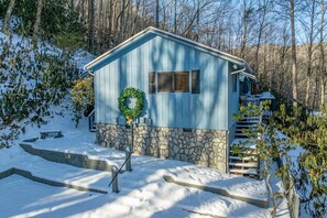 A small, single-story cabin with a stone foundation and a blue exterior, surrounded by snow-covered ground and trees.