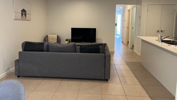 A modern living room with a grey sofa, a flat-screen TV, and a small side table. A hallway leads to a kitchen area with a sink and a countertop. - Stylish Retreat in Macquarie Links - Golf, Comfort & Tranquility (Macquarie Links)