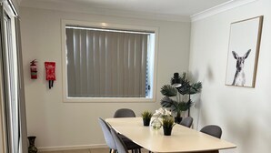 A dining area with a table and chairs, a window with blinds, a fire extinguisher, and a framed picture on the wall. - Stylish Retreat in Macquarie Links - Golf, Comfort & Tranquility (Macquarie Links)