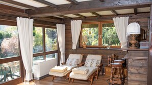 L’intérieur d’un chalet en bois, avec de grandes fenêtres, deux fauteuils rembourrés, un tabouret en bois et un radiateur.