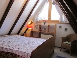 A bedroom with a bed, a nightstand, a lamp, a chair, a window with curtains, and a shelf with books and a plant.
