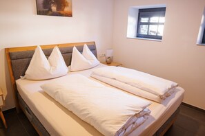 A neatly made bed with white linens and pillows, a bedside table with a lamp, and a window with blinds.