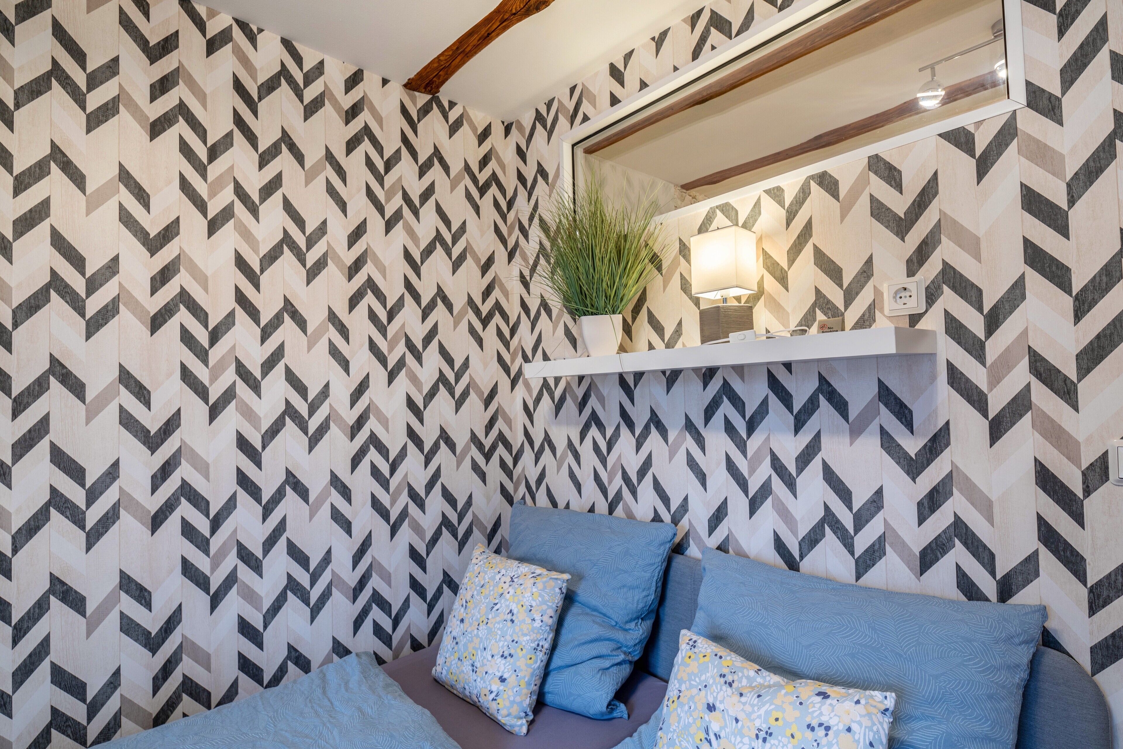 A bedroom with a chevron-patterned wallpaper, a bed with blue and floral pillows, a bedside table with a lamp, and a potted plant on a shelf.