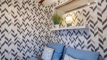 A bedroom with a chevron-patterned wallpaper, a bed with blue and floral pillows, a bedside table with a lamp, and a potted plant on a shelf.