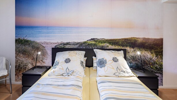 A double bed with floral pillows, a nightstand with a lamp, and a large beach scene mural on the wall. - Apartment "Waldeckchen" with Private Terrace & Wi-Fi (Waldeck)