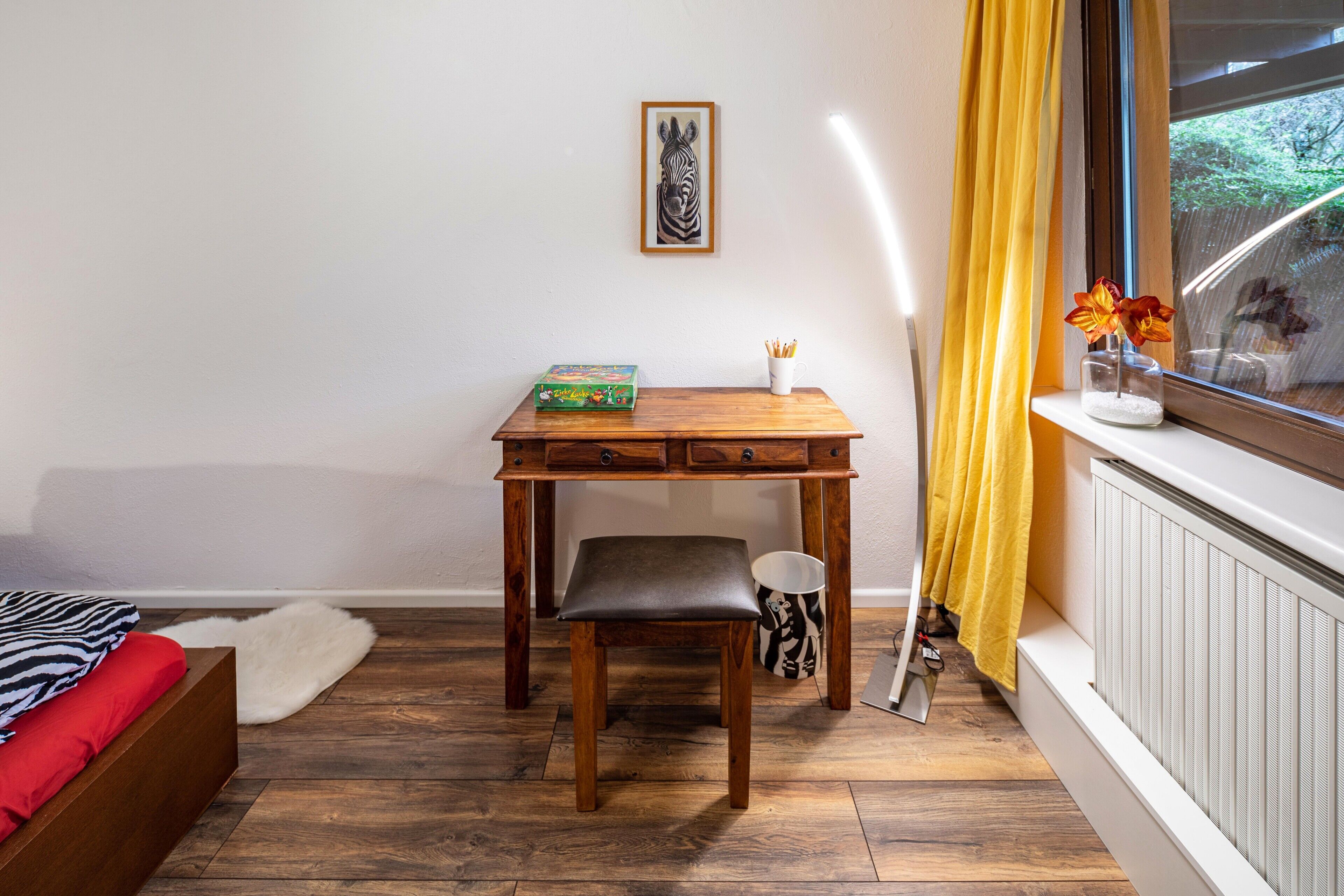 Una habitación con escritorio de madera, una silla, una cama con ropa de cama roja y negra, una obra de arte enmarco y una ventana con vistas a zonas verdes.