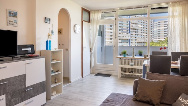 Ett modernt vardagsrum med en platt-TV, en soffa, en matplats och utsikt över höga byggnader.