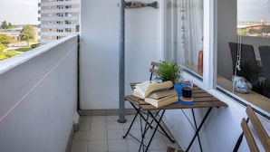 En balkong med ett bord, en stol, en växt och ett fönster med utsikt över byggnader.