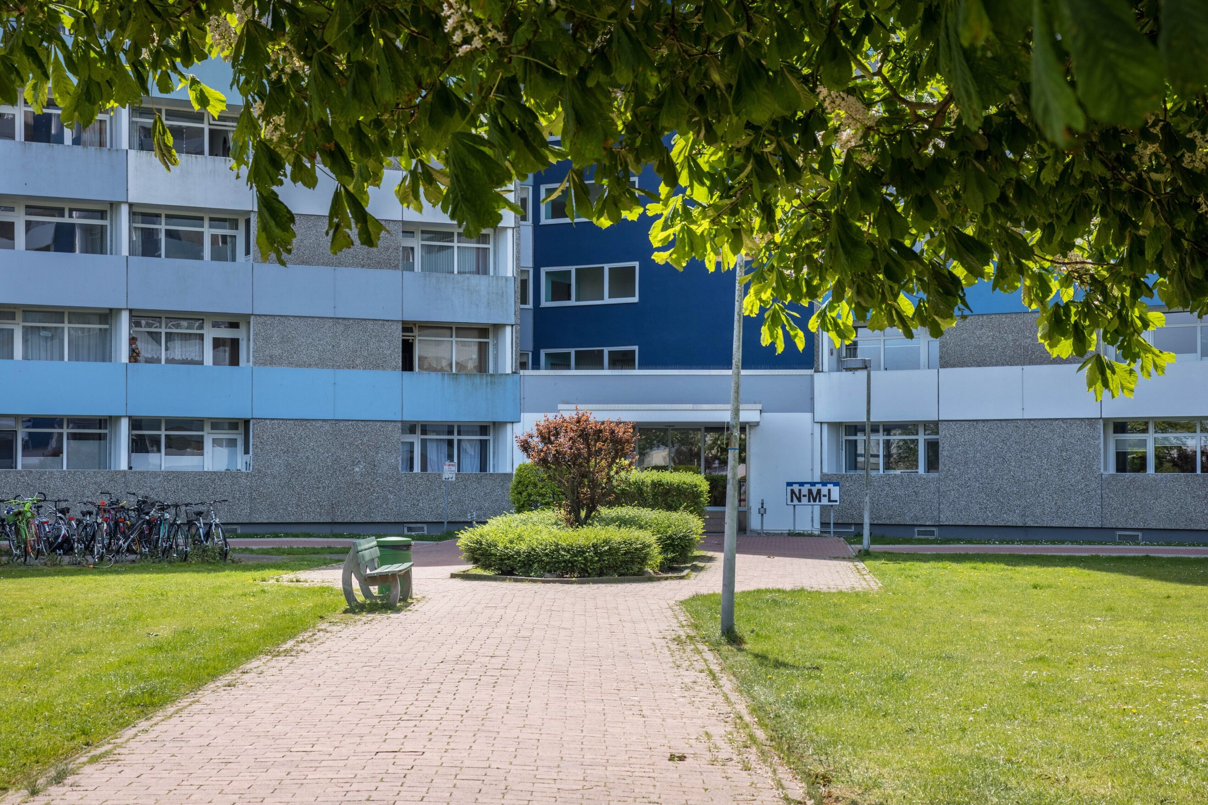 Un bâtiment à la façade bleue et grise, un chemin pavé et un banc.