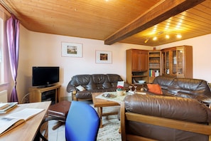 A living room with a leather sofa, a television, a wooden cabinet, and a table with books and a vase.