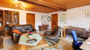 Ein gemütliches Wohnzimmer mit Holzbalkendecke, Ledermöbeln, einem Couchtisch mit Büchern und einem Bett mit einem hölzernen Kopfteil.
