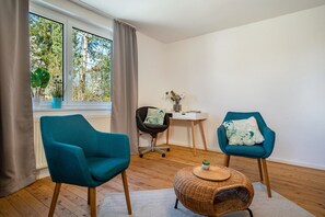 A room with two teal armchairs, a small round table with a plant, a black office chair, a white side table, and a view of trees through a window.