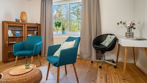 Una habitación con piso de madera, dos sillones azul verdoso, una silla de oficina negra, un escritorio blanco, una mesita de mimbre, una estantería y una ventana con cortinas.