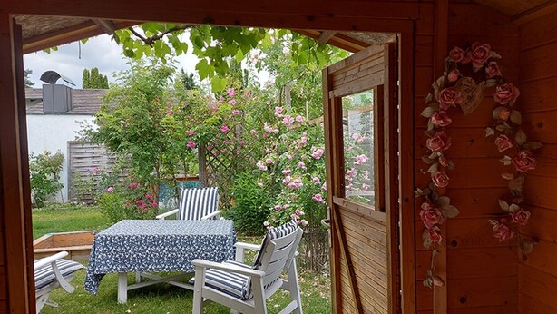En öppen träkonstruktion med ett bord och stolar, en trädgård med rosa blommor och en trävägg med ett blommönster.