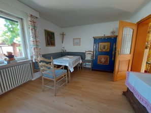 Ein Zimmer mit Esstisch, Stühlen, Heizkörper, einem Fenster mit Vorhängen, einer Tür und einem Kruzifix an der Wand.