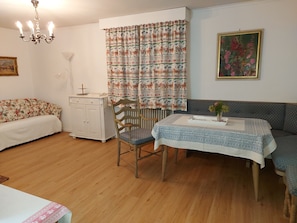 Ein Zimmer mit Holzboden, einem Esstisch mit gemustertem Tischlein, einer Couch, einem Schrank und einer Heizung.