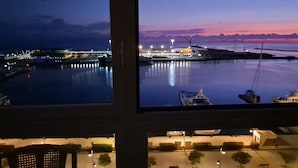 A marina at dusk with boats docked, a bridge in the background, and a view of a building with a flat roof and a small parking area with trees.