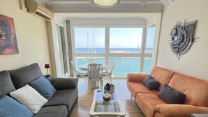 A living room with a grey sofa, an orange sofa, and a white coffee table. There is a view of the sea and boats through large windows.