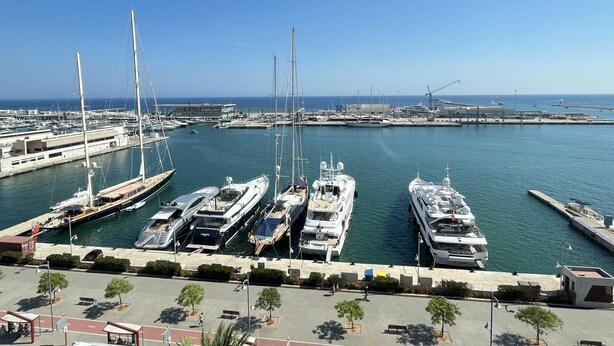 A marina with multiple yachts docked, a clear sky, and a calm sea.